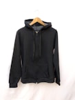 A black hoodie is displayed on a wooden hanger. The hoodie has a zipper in the front, two front pockets, and drawstrings for the hood. The background is plain white.