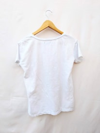 A plain white t-shirt is displayed on a wooden hanger against a light background. The t-shirt is short-sleeved and appears to be made of a lightweight material.