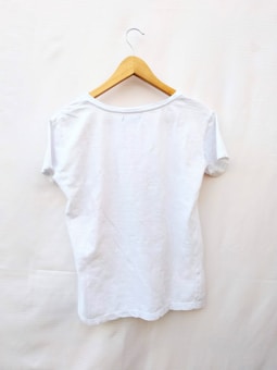 A plain white t-shirt is displayed on a wooden hanger against a light background. The t-shirt is short-sleeved and appears to be made of a lightweight material.