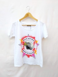 A white t-shirt hanging on a wooden hanger features a graphic design. The center showcases an eagle's head within a star shape, surrounded by flames and text that reads 'Rock and Roll' and 'Made in Heaven.'