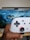 A hand is holding a white gaming controller with black buttons and an orange accent around the joystick. In the background, a television screen displays a scenic image featuring clouds and mountains.