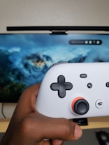 A hand is holding a white gaming controller with black buttons and an orange accent around the joystick. In the background, a television screen displays a scenic image featuring clouds and mountains.
