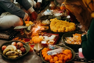 A group of people engaged in a traditional ceremony or ritual, with various offerings including fruits, sweets, and marigold flowers arranged on plates. A small oil lamp is lit, casting a warm glow over the scene. The participants are dressed in formal or festive attire and appear to be seated on a decorated floor.