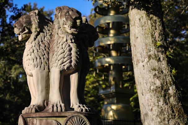 A detailed sculpture of three lion heads with ornate manes stands prominently. The background features a tall golden pagoda and lush green trees, highlighting a serene outdoor setting.