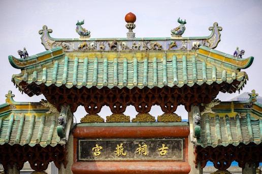 An ornate traditional Chinese roof with intricate carvings and decorations, featuring green tiles and sculptures of mythical creatures. The structure is adorned with yellow and gold accents and contains Chinese characters in the center.