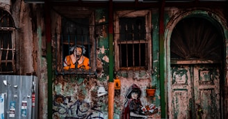 An aged, graffiti-covered wall features a painted figure in a striped prison outfit looking out from a barred window. Below, a mural of a woman in a dark cloak and red hood is holding a spray can. The wall and door are weathered, with peeling green and brown paint, surrounded by various metal elements and pots.