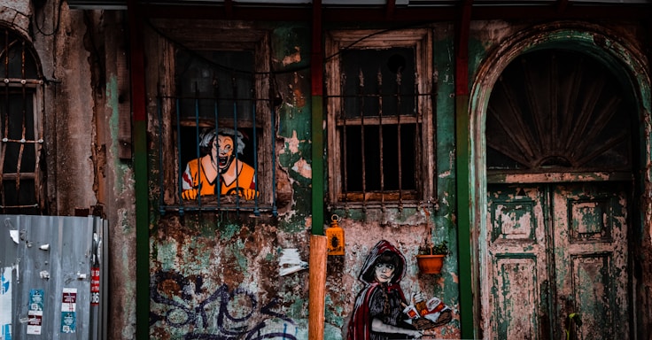 An aged, graffiti-covered wall features a painted figure in a striped prison outfit looking out from a barred window. Below, a mural of a woman in a dark cloak and red hood is holding a spray can. The wall and door are weathered, with peeling green and brown paint, surrounded by various metal elements and pots.