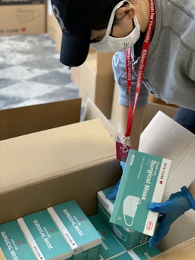 A person wearing gloves, a cap, and a face mask is packing boxes containing surgical masks. The individual is handling a box of 'BYD CARE' labeled surgical masks, placing them into a larger cardboard box.