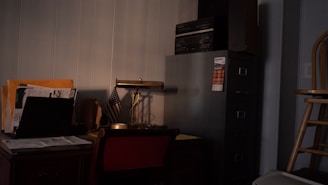 A dimly lit office corner featuring a wooden desk cluttered with paperwork, a laptop, and an American flag. A banker’s lamp sits on the desk, alongside a filing cabinet topped with an audio stereo system. A wooden chair is partially visible in the foreground.