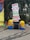 Two Minion figures are placed beside a directional signboard. The signboard includes words such as Propulsion, Robo Soccer, Technostunt, Career Guidance, Entrepreneurship, Creed Cultural, and Click a Pic, in various vibrant colors. The background features green foliage and a large colorful object on the left.