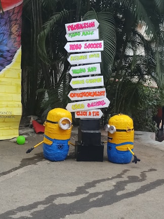 Two Minion figures are placed beside a directional signboard. The signboard includes words such as Propulsion, Robo Soccer, Technostunt, Career Guidance, Entrepreneurship, Creed Cultural, and Click a Pic, in various vibrant colors. The background features green foliage and a large colorful object on the left.