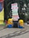 Two Minion figures are placed beside a directional signboard. The signboard includes words such as Propulsion, Robo Soccer, Technostunt, Career Guidance, Entrepreneurship, Creed Cultural, and Click a Pic, in various vibrant colors. The background features green foliage and a large colorful object on the left.