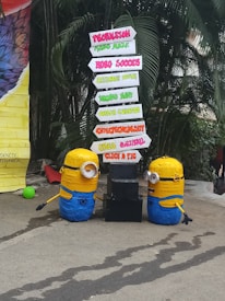 Two Minion figures are placed beside a directional signboard. The signboard includes words such as Propulsion, Robo Soccer, Technostunt, Career Guidance, Entrepreneurship, Creed Cultural, and Click a Pic, in various vibrant colors. The background features green foliage and a large colorful object on the left.