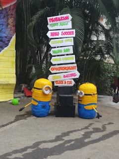 Two Minion figures are placed beside a directional signboard. The signboard includes words such as Propulsion, Robo Soccer, Technostunt, Career Guidance, Entrepreneurship, Creed Cultural, and Click a Pic, in various vibrant colors. The background features green foliage and a large colorful object on the left.