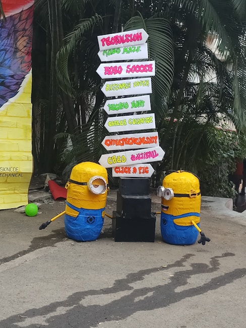Two Minion figures are placed beside a directional signboard. The signboard includes words such as Propulsion, Robo Soccer, Technostunt, Career Guidance, Entrepreneurship, Creed Cultural, and Click a Pic, in various vibrant colors. The background features green foliage and a large colorful object on the left.