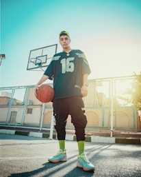 A young person stands on a basketball court holding a basketball. They are wearing athletic clothing, including a jersey and colorful sneakers. A basketball hoop is visible in the background with bright sunlight streaming in.