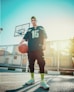 A young person stands on a basketball court holding a basketball. They are wearing athletic clothing, including a jersey and colorful sneakers. A basketball hoop is visible in the background with bright sunlight streaming in.