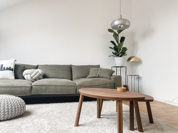 A modern living room features a sage green sofa adorned with two pillows, one of which is uniquely knotted. A sleek wooden coffee table stands on a plush light beige rug. A potted plant in a white pot is placed on a thin black metal stand, complemented by a metallic lamp with a gold finish. The ambience is enhanced by a minimalist overhead lamp, with the walls painted in a soft white hue.