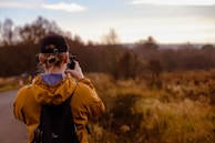 A person wearing a brown jacket, black cap, and a backpack is taking a photo of a natural landscape. The area around them is filled with autumn-colored foliage, with trees in the distance under a partly cloudy sky.