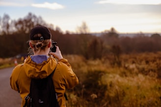 A person wearing a brown jacket, black cap, and a backpack is taking a photo of a natural landscape. The area around them is filled with autumn-colored foliage, with trees in the distance under a partly cloudy sky.