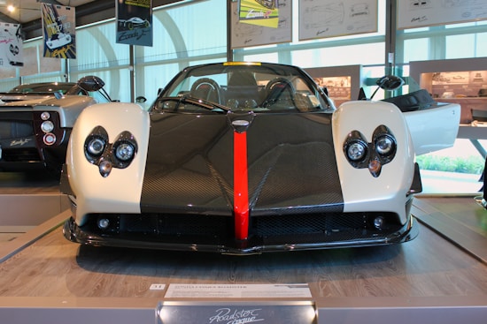 A sleek, modern sports car with a distinct design, featuring a carbon fiber hood and white body panels. The front grille has circular headlights and a prominent red stripe running down the center. The surrounding environment suggests an indoor showroom with other cars and automotive posters visible.