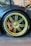 A close-up of a luxury car's wheel with a golden-colored rim and large brake calipers. The wheel is mounted with Pirelli tires, and the brake caliper is painted red. The surrounding area includes a carbon fiber fender with a glossy finish, and the setting appears to be indoors with framed pictures visible on the wall.