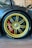 A close-up of a luxury car's wheel with a golden-colored rim and large brake calipers. The wheel is mounted with Pirelli tires, and the brake caliper is painted red. The surrounding area includes a carbon fiber fender with a glossy finish, and the setting appears to be indoors with framed pictures visible on the wall.