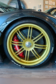 A close-up of a luxury car's wheel with a golden-colored rim and large brake calipers. The wheel is mounted with Pirelli tires, and the brake caliper is painted red. The surrounding area includes a carbon fiber fender with a glossy finish, and the setting appears to be indoors with framed pictures visible on the wall.