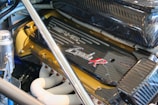 A close-up of a high-performance engine featuring a carbon fiber cover with branding and text. The polished metal surfaces and white pipes reflect precision engineering and advanced automotive design. Various components such as bolts, tubes, and metallic fittings are visible, highlighting the complexity and functionality of the engine.