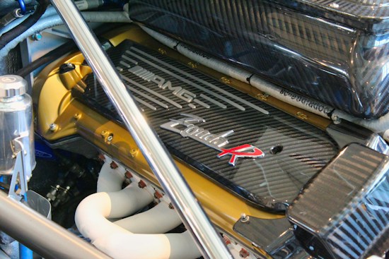 A close-up of a high-performance engine featuring a carbon fiber cover with branding and text. The polished metal surfaces and white pipes reflect precision engineering and advanced automotive design. Various components such as bolts, tubes, and metallic fittings are visible, highlighting the complexity and functionality of the engine.