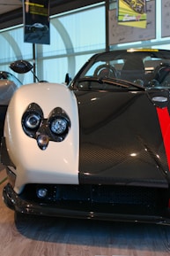A close-up view of a car's front, with a sleek, sporty design and a distinctive headlight arrangement. The car's body features a contrasting color scheme, combining black carbon fiber with a white panel.