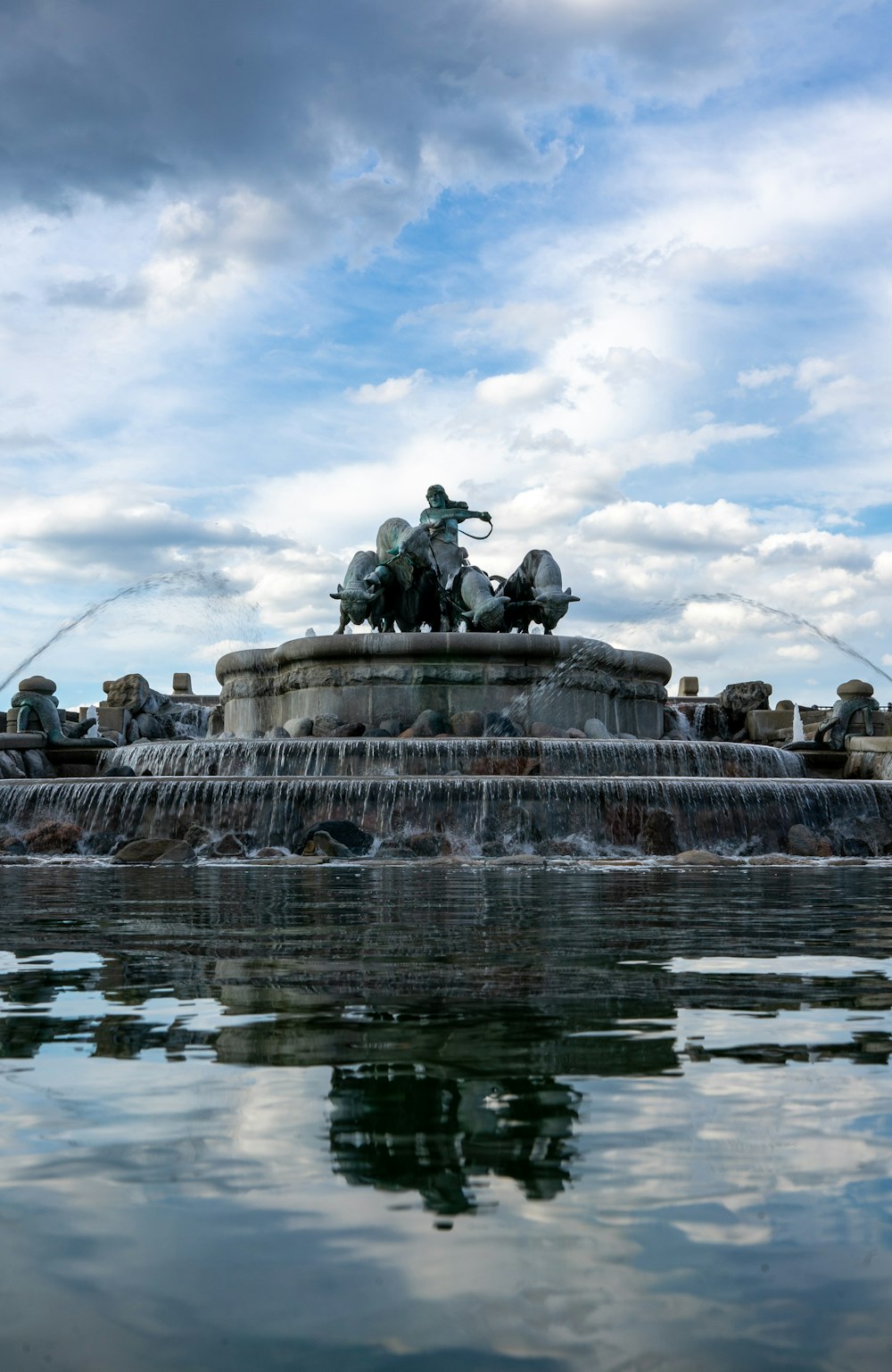 people sitting on bench statue on water fountain under white clouds and blue sky during daytime