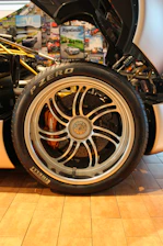 A detailed close-up of a car's wheel and tire is surrounded by various automotive-themed posters. The tire is branded Pirelli P Zero, and the alloy wheel features intricate spoke designs with visible brake components underneath.