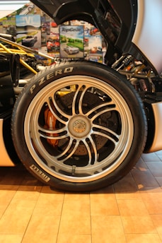 A detailed close-up of a car's wheel and tire is surrounded by various automotive-themed posters. The tire is branded Pirelli P Zero, and the alloy wheel features intricate spoke designs with visible brake components underneath.
