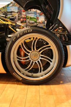 A detailed close-up of a car's wheel and tire is surrounded by various automotive-themed posters. The tire is branded Pirelli P Zero, and the alloy wheel features intricate spoke designs with visible brake components underneath.