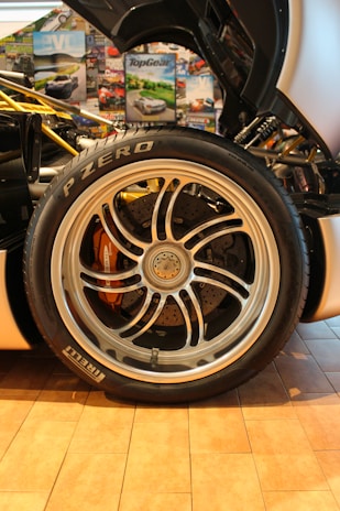 A detailed close-up of a car's wheel and tire is surrounded by various automotive-themed posters. The tire is branded Pirelli P Zero, and the alloy wheel features intricate spoke designs with visible brake components underneath.