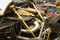 A close-up view of a car's engine compartment showcasing its intricate design and engineering. The image highlights carbon fiber components, metallic structures, and a yellow cover over part of the engine. Various automotive magazines, including titles like Top Gear and Autocar, form the background.