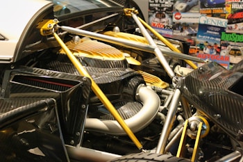 A close-up view of a car's engine compartment showcasing its intricate design and engineering. The image highlights carbon fiber components, metallic structures, and a yellow cover over part of the engine. Various automotive magazines, including titles like Top Gear and Autocar, form the background.
