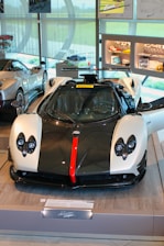 A luxury sports car is displayed in a showroom with its doors open. The car features a sleek design with a black and white color scheme, accented by a bold red stripe down the center of the front. The interior of the showroom includes other high-end cars and various automotive displays and memorabilia on the walls and shelves.