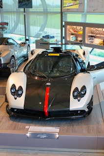 A luxury sports car is displayed in a showroom with its doors open. The car features a sleek design with a black and white color scheme, accented by a bold red stripe down the center of the front. The interior of the showroom includes other high-end cars and various automotive displays and memorabilia on the walls and shelves.