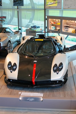 A luxury sports car is displayed in a showroom with its doors open. The car features a sleek design with a black and white color scheme, accented by a bold red stripe down the center of the front. The interior of the showroom includes other high-end cars and various automotive displays and memorabilia on the walls and shelves.