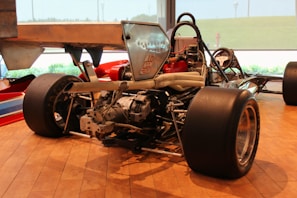A vintage race car is displayed indoors, with its rear components such as the large tires, engine, and exhaust system clearly visible. The polished wooden flooring and large windows in the background suggest a showroom or museum setting.