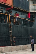 Several people are standing on the deck of a large ship next to stacked shipping containers. They are looking down at a man on the dock, who is gesturing upwards. A rope ladder hangs alongside the ship's hull.