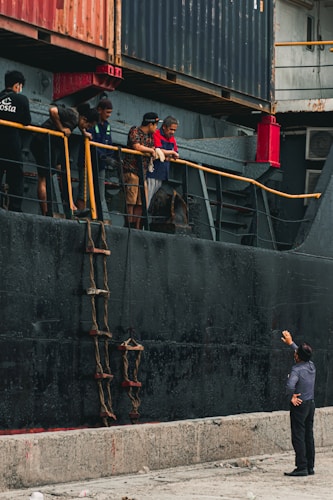 Several people are standing on the deck of a large ship next to stacked shipping containers. They are looking down at a man on the dock, who is gesturing upwards. A rope ladder hangs alongside the ship's hull.
