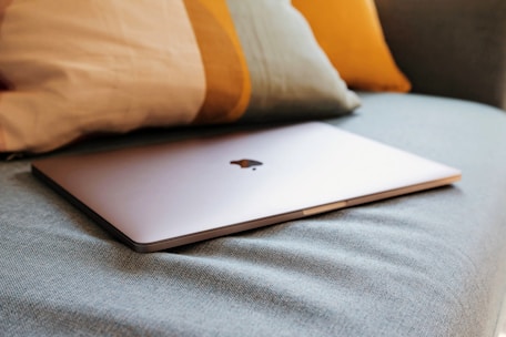 A sleek, closed laptop with a metallic finish is resting on a soft, textured fabric surface, accompanied by cushions in the background featuring muted colors.
