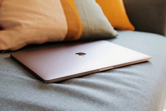 A sleek, closed laptop with a metallic finish is resting on a soft, textured fabric surface, accompanied by cushions in the background featuring muted colors.