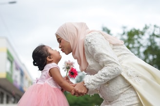 A woman in an elegant white dress and a light pink hijab is bending forward, holding hands with a young girl in a pink dress. They are sharing a playful and affectionate moment, with the woman puckering her lips towards the girl. The girl holds a small bouquet of flowers, and the background features trees and a building on the side.