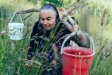 An elderly woman with a headscarf is carrying two buckets attached to a wooden yoke across her shoulders, walking through a field of tall grass. The scene appears to be rural and captures a moment of hard work and resilience.