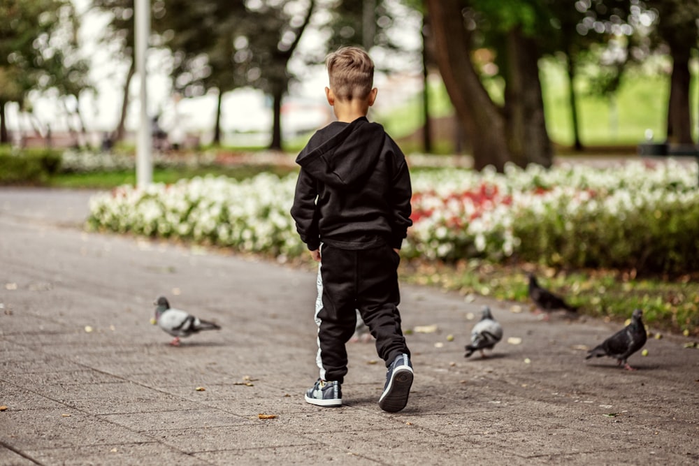 boy in black jacket and black pants walking on gray concrete pavement with pigeons during daytime