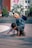 Two children are playing outdoors on a paved surface. One child wearing a polka dot scarf is sitting on the back of the other child, who is crawling on the ground. There is a background of steps, trees, a building with colorful wall art, and other people in the distance.